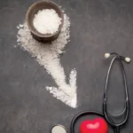 "ملعقة ملح فوق طاولة مع أطعمة يومية، يوضح أضرار زيادة الملح في الغذاء على ضغط الدم والصحة العامة."