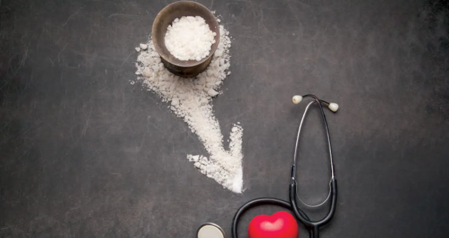 "ملعقة ملح فوق طاولة مع أطعمة يومية، يوضح أضرار زيادة الملح في الغذاء على ضغط الدم والصحة العامة."